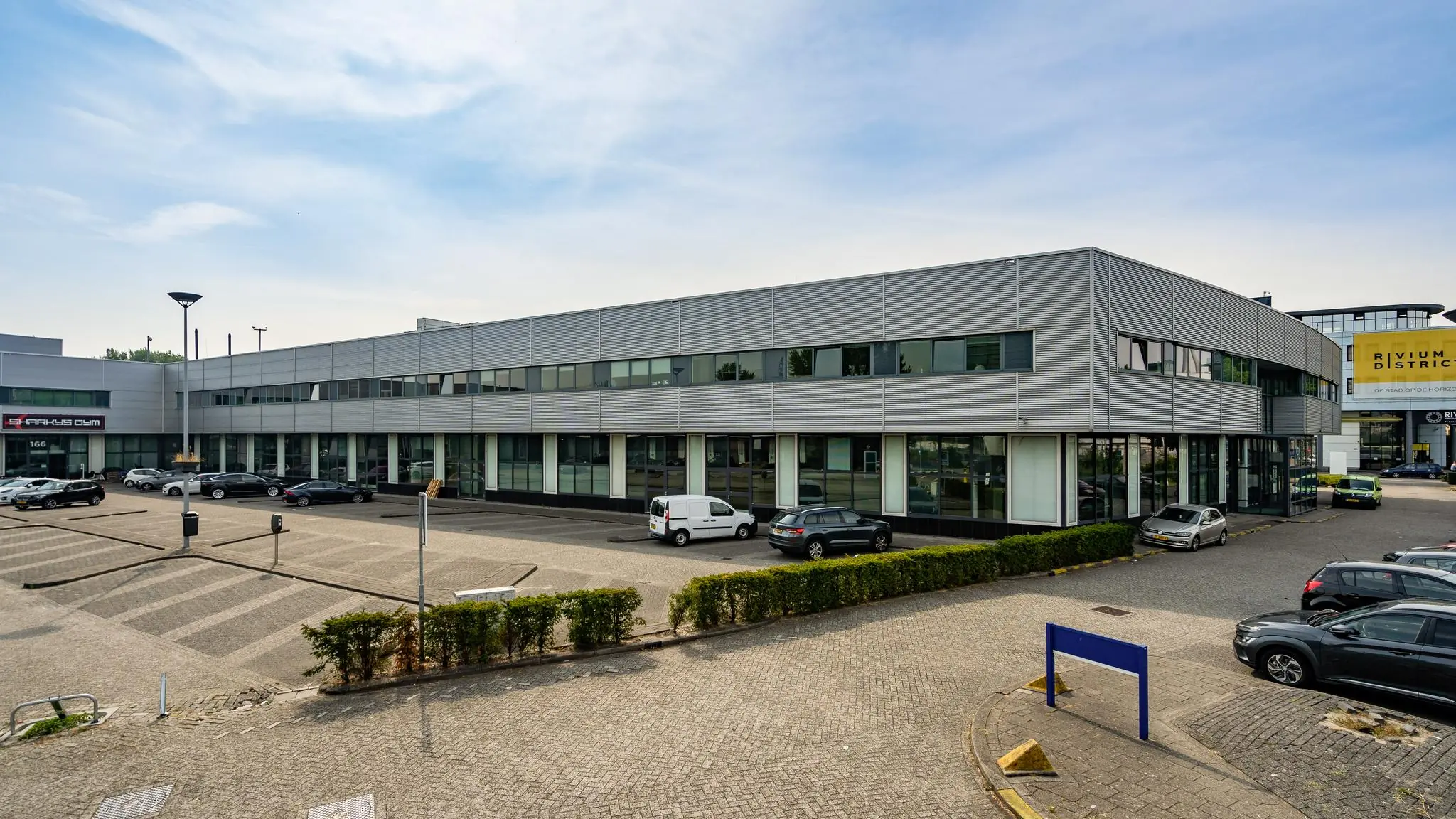 Grijs kantoorgebouw aan de Rivium Boulevard met parkeerplaatsen en diverse geparkeerde auto's.